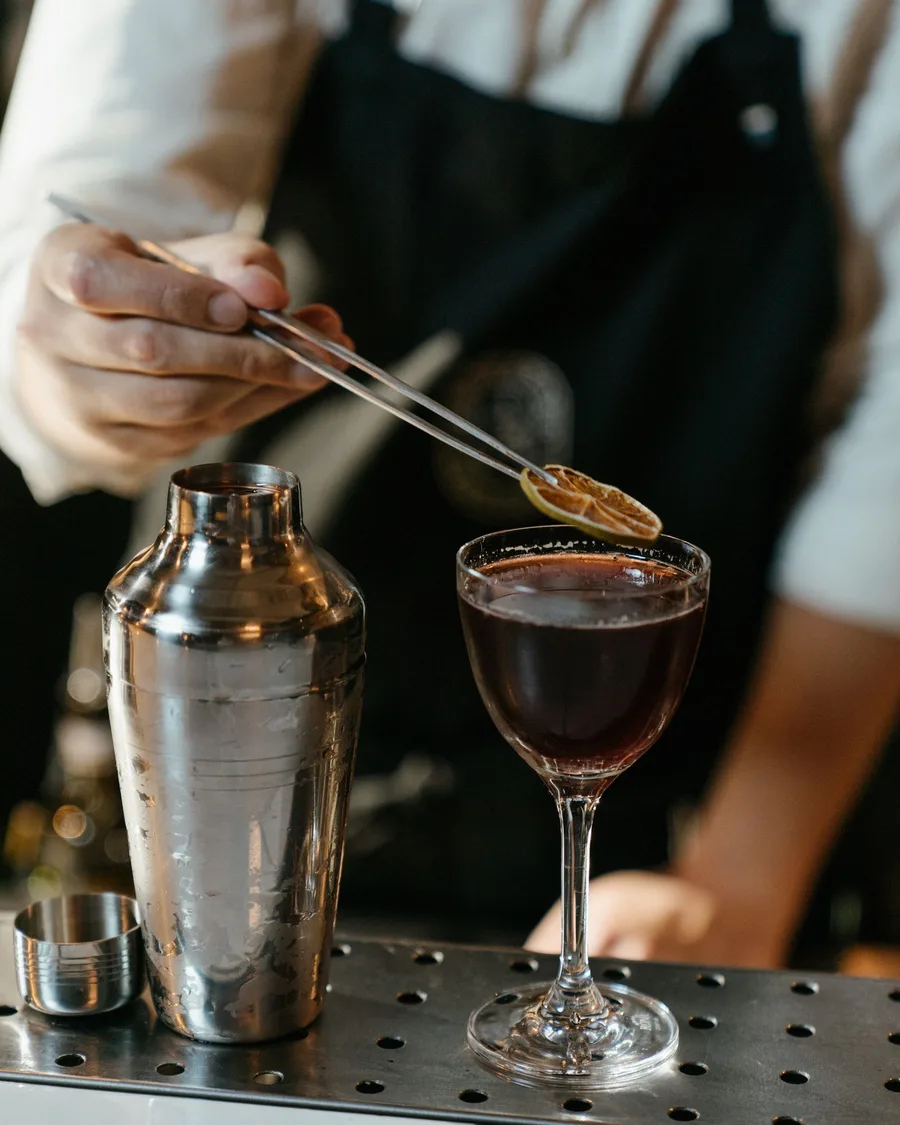 A bartender garnishing a dark coupe cocktail with a dried citrus wheel, a shaker beside it on the bar