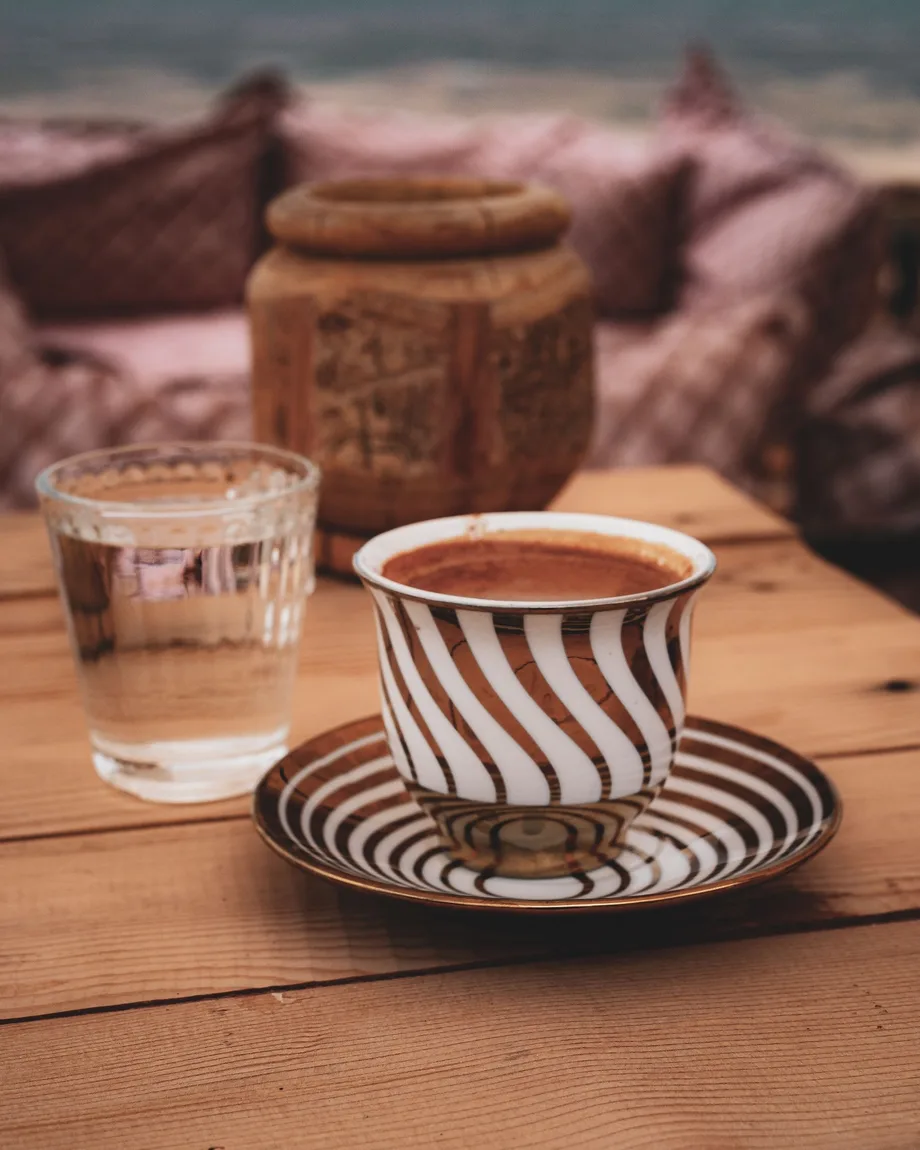 A striped Turkish coffee cup and a glass of water on a rustic wooden cafe table by the sea