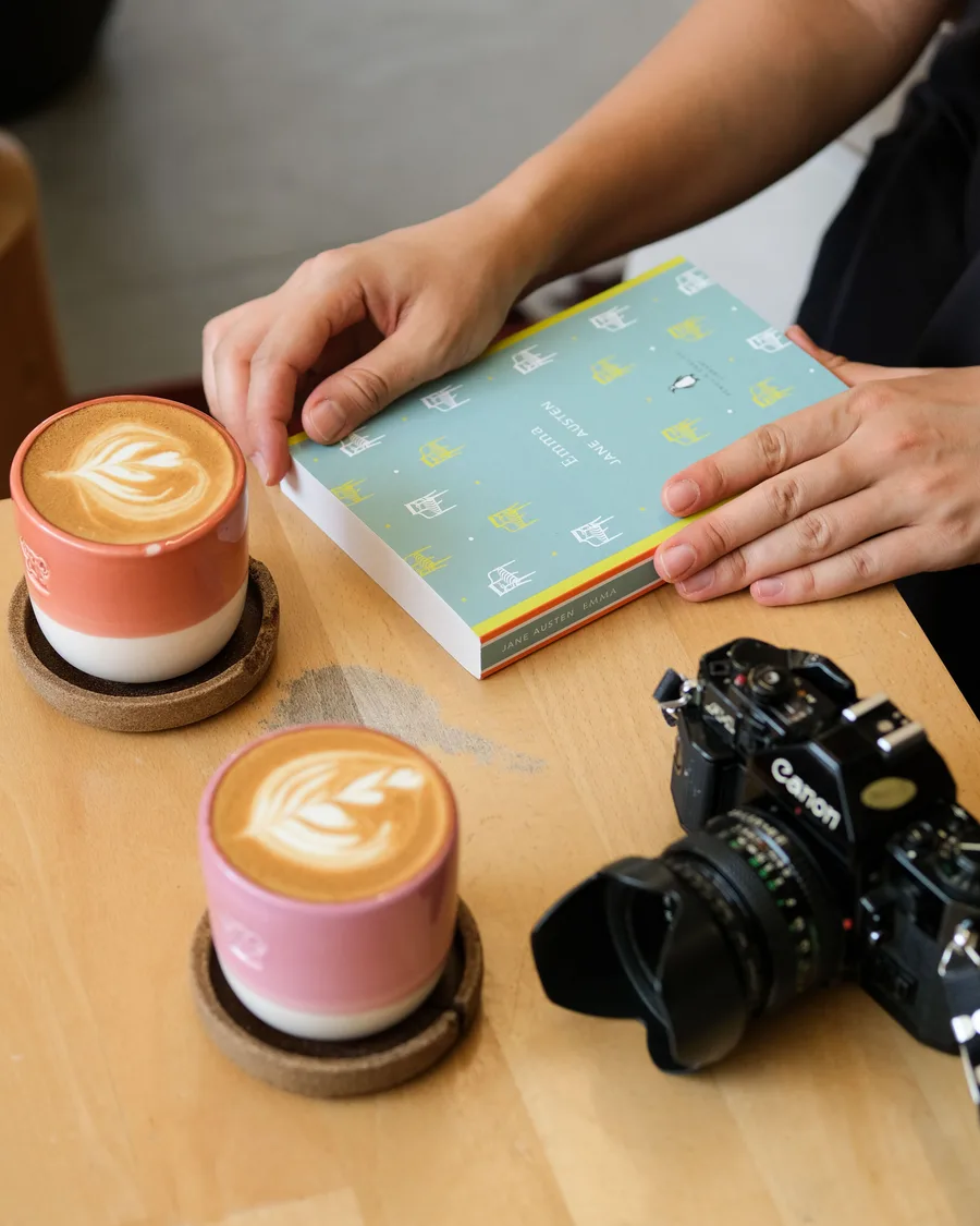 Two lattes with latte art beside a book and a film camera on a cafe table