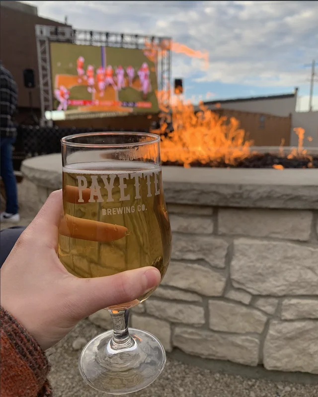 A hand holding a Payette Brewing Co. stemmed pour glass in front of a stone fire pit