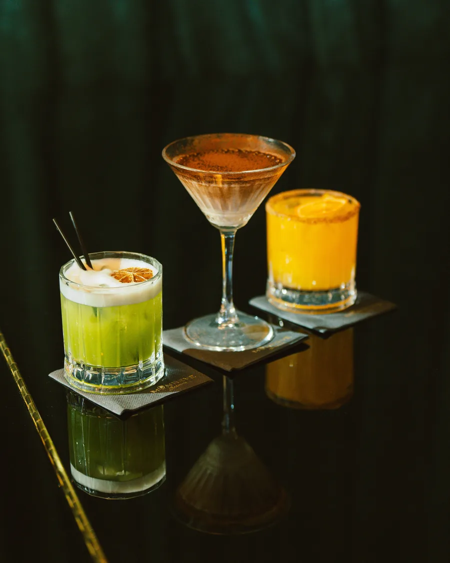Three cocktails, a green sour, a cocoa martini and an orange margarita, on a dark reflective bar