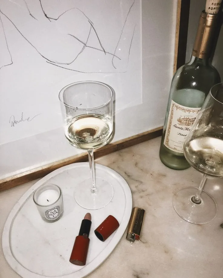 A glass of white wine on a marble tray beside a bottle of Pinot, a candle and a lipstick